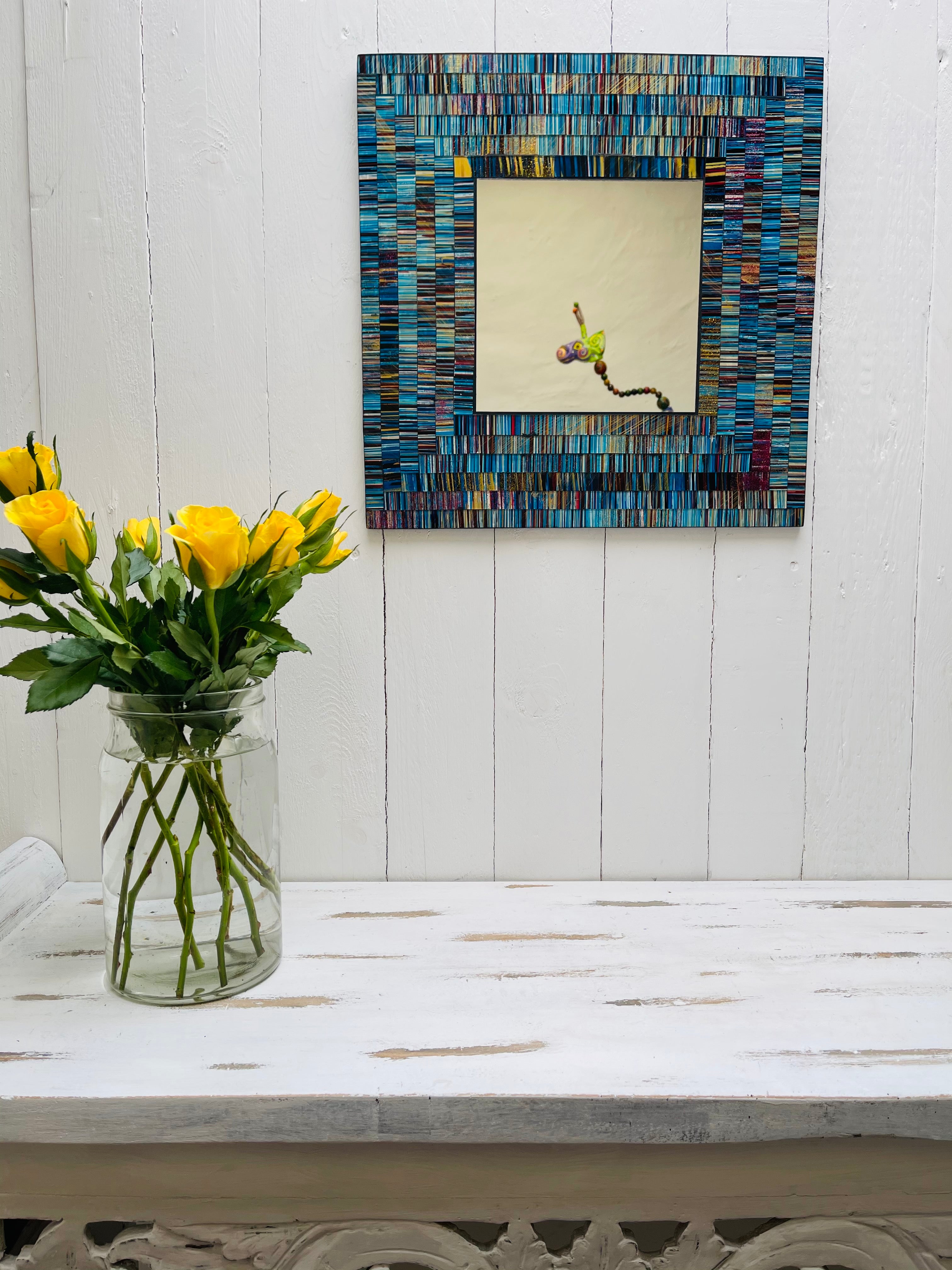 display front view of mosaic square mirror hung on the wall with vase of roses below on table